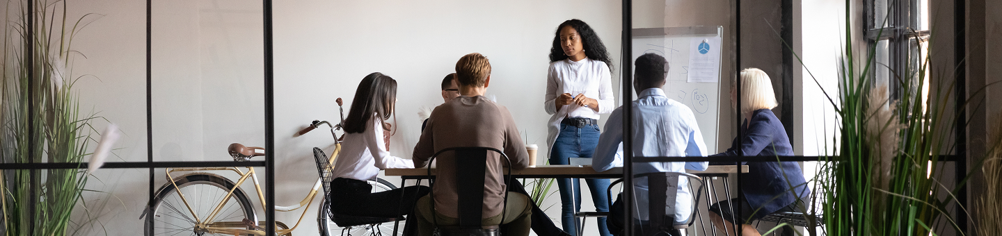 People standing and sitting around a table, having a meeting, with a dry erase board and bicycle in the background